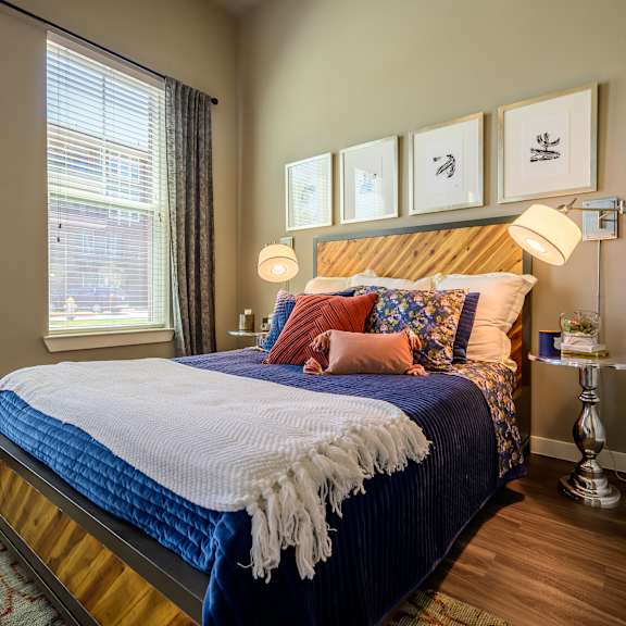 here at harmony luxury, this stylish bedroom features a warm wood bed frame, deep blue bedding layered with patterned and blush accent pillows, modern wall-mounted reading lights, and soft natural light from large windows that create a cozy, inviting retreat.