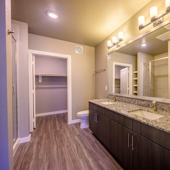 here at harmony luxury, this spacious bathroom features a modern double-sink vanity with a granite countertop, dark wood cabinetry, warm overhead lighting, wood-style flooring, and a large walk-in shower, creating a clean and polished everyday space.