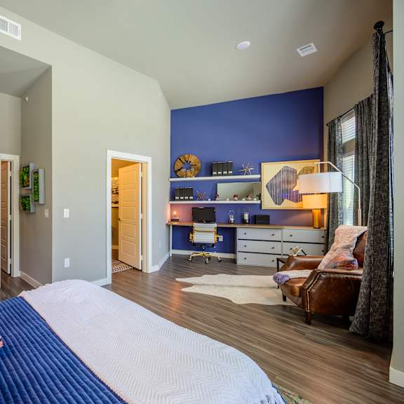 a spacious bedroom here at harmony luxury with warm wood-style flooring, a bold blue accent wall, and a dedicated workspace featuring built-in shelving and a desk, complemented by soft natural light, modern décor, and a comfortable seating area that creates a stylish, inviting atmosphere.