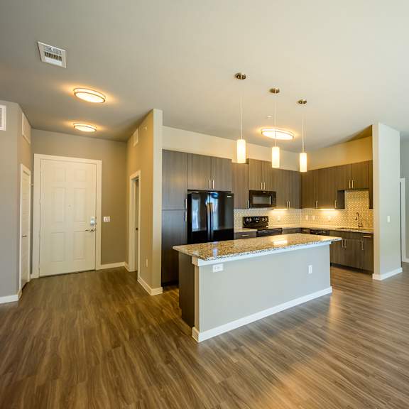 a spacious, open-concept kitchen here at harmony luxury with warm wood-style flooring, dark modern cabinets, granite countertops, and a large island with bar-style seating, complemented by stainless steel appliances, pendant lighting, and a bright, contemporary feel.