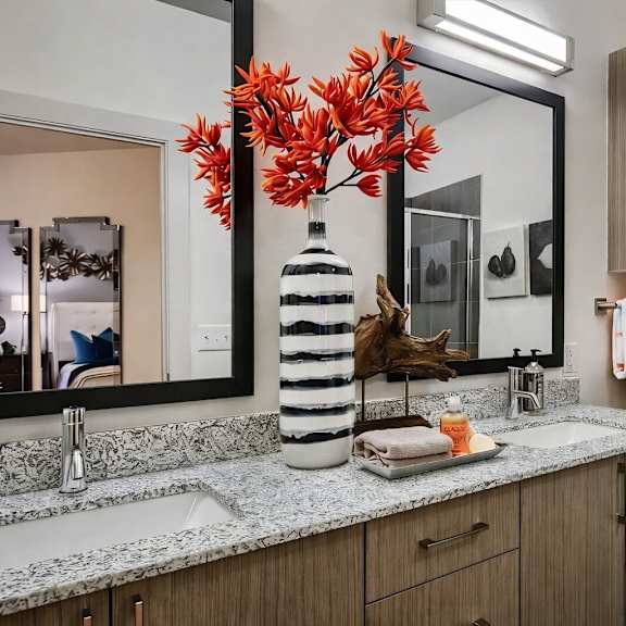 a bathroom here at skyline farmers market with dual sinks, speckled granite vanity, light wood cabinets, black framed mirrors, and standout orange towels with matching orange floral décor on the counter.