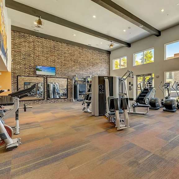 a fitness center here at skyline farmers market with large windows, brick accent wall, treadmills and ellipticals, strength machines, mirrors, and high ceilings with exposed beams and bright natural light.
