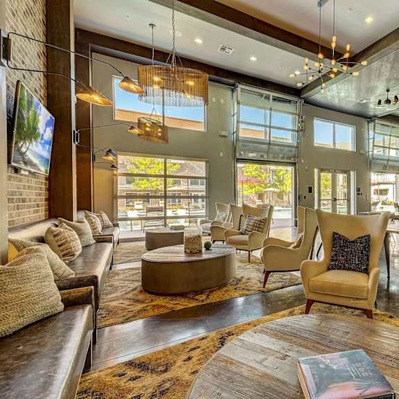 a resident clubhouse lounge here at skyline farmers market with tan accent chairs, brown leather sofa, round wood tables, warm chandeliers, exposed beams, and large glass garage doors bringing in bright natural light.