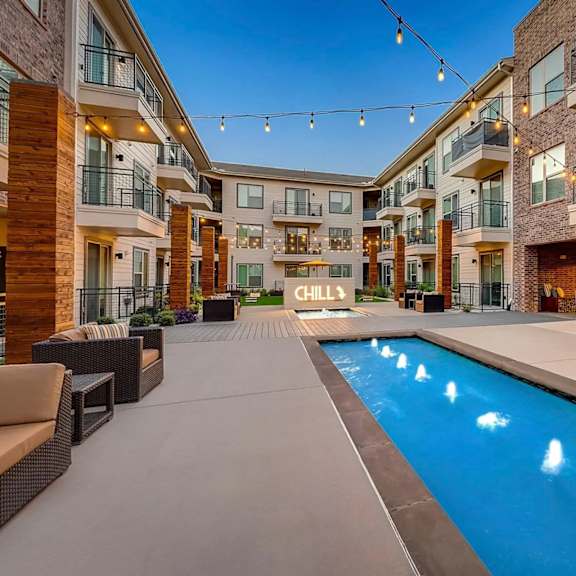 a courtyard pool here at skyline farmers market with a narrow blue lap pool, glowing string lights, wood columns, yellow umbrella, and modern patio seating creating a warm, social outdoor space at dusk.