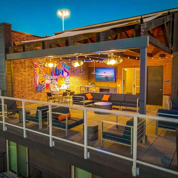 a rooftop lounge here at skyline farmers market with dark seating and standout orange accent pillows, covered pergola, string lights, mural wall, and open-air city-view terrace at dusk.