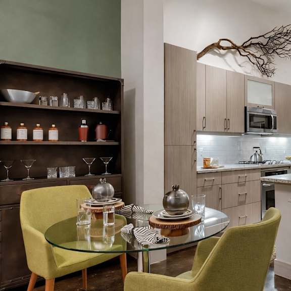 a dining and kitchen area here at skyline farmers market with light wood cabinets, stainless appliances, a white island, glass round table, and standout green dining chairs beside a dark wood display shelf.