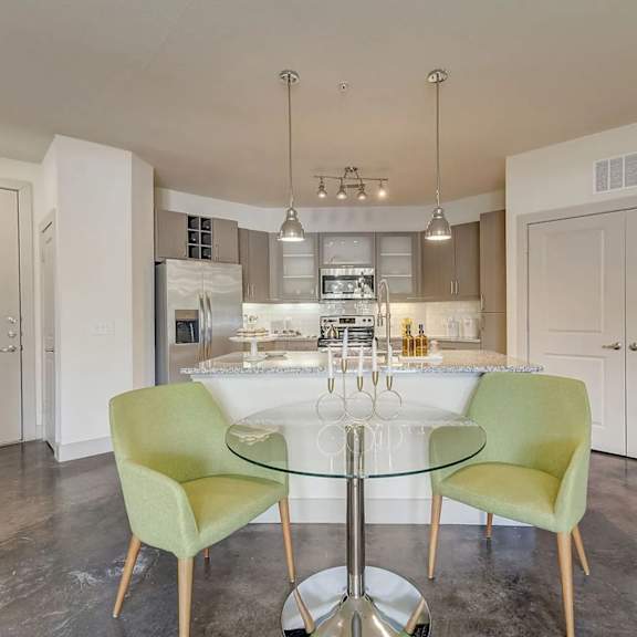 a dining nook here at skyline farmers market with a round glass table, two standout green accent chairs, pendant lights, and an open kitchen with granite island and stainless appliances behind it.