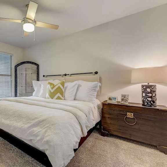 a bedroom here at skyline farmers market with a white bed, yellow chevron accent pillow, dark wood nightstand, patterned lamp, beige carpet, ceiling fan, wall art, and a small window bringing in soft natural light.