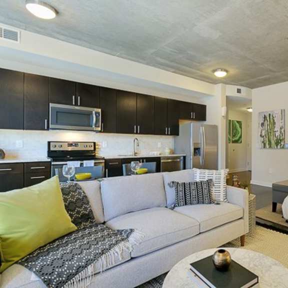 a modern open studio layout here at victory place with a full kitchen, dark cabinets, stainless steel appliances, a cozy sofa, and a sleeping area with green accent bedding and city-view windows.