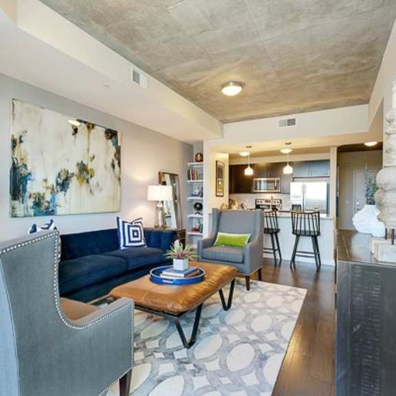 a modern living room here at victory place with dark wood flooring, a blue sofa, accent chairs, abstract wall art, a coffee table, and an open view into the kitchen with bar seating and pendant lights.