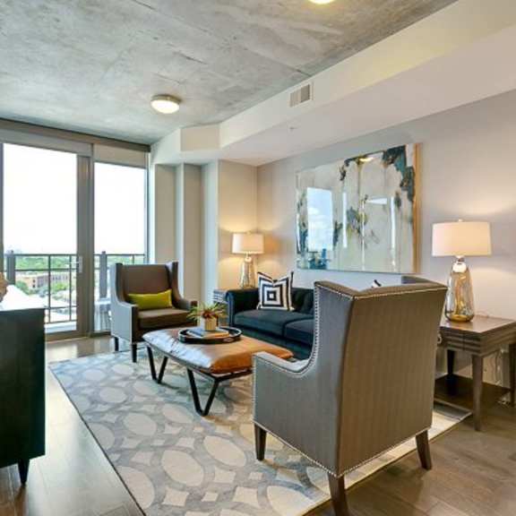 a bright living room here at victory place with dark wood flooring, a blue sofa, accent chairs, table lamps, abstract wall art, and a private balcony view through large glass doors.