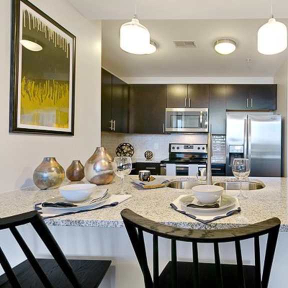 a modern kitchen island here at victory place with granite-style countertops, pendant lighting, bar seating for four, dark cabinets, and stainless steel appliances in an open-concept layout.
