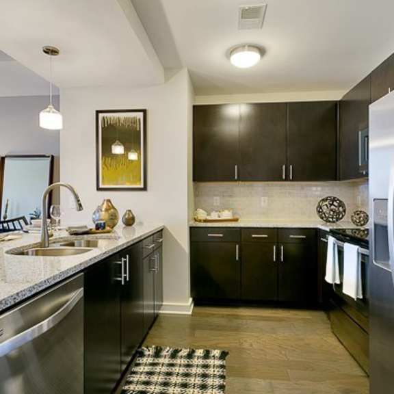 a modern l-shaped kitchen here at victory place with dark cabinets, stainless steel appliances, granite-style countertops, pendant lighting, and a double sink with bar seating along the island.