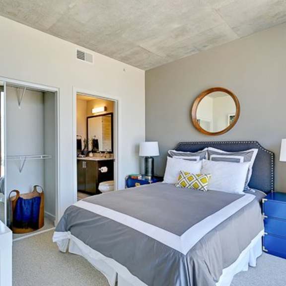 a bedroom here at victory place, including a nighstand, modern lamp, a mirror above the bed, with a sliding  closet in the wall. the space is encompassed by bright natural light