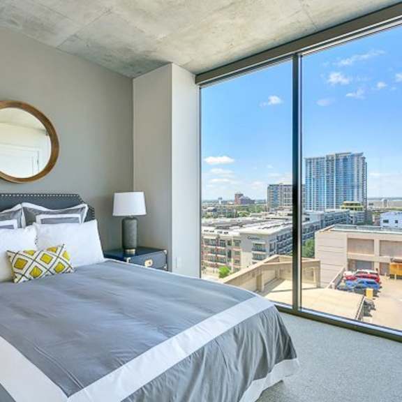 a bedroom here at victory place encompassed by  a large window covering one side of the wall, bringing in natural light. above the bed sits a modern rounded mirror adding depth and coziness to the space.
