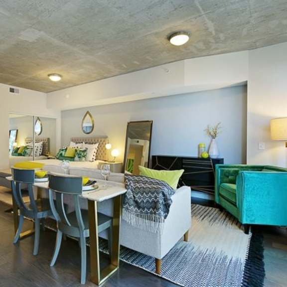 a modern open studio here at victory place with dark wood flooring, a dining table, cozy seating, green accent furniture, and a sleeping area with decorative bedding and nearby bathroom access.