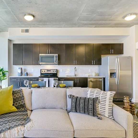 a modern open kitchen and living area here at victory place with dark cabinets, stainless steel appliances, a tile backsplash, a cozy sofa with accent pillows, and large windows with city views.