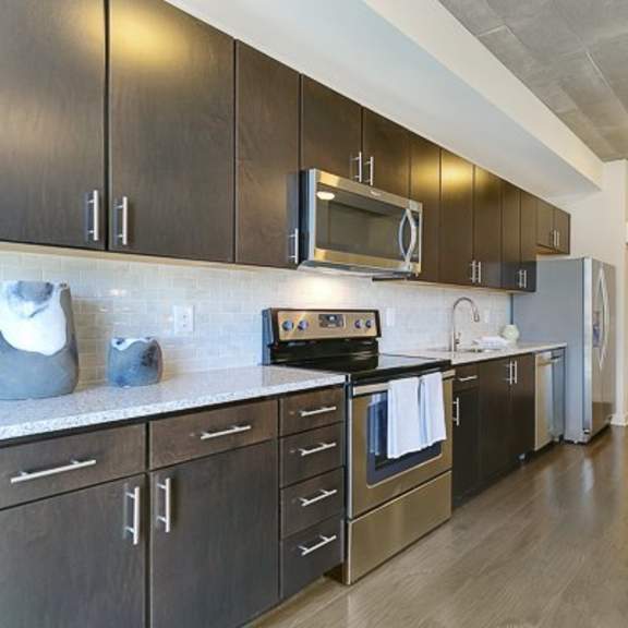 a sleek galley-style kitchen here at victory place with dark wood cabinets, stainless steel appliances, a tile backsplash, modern countertops, and a nearby dining table set for four.
