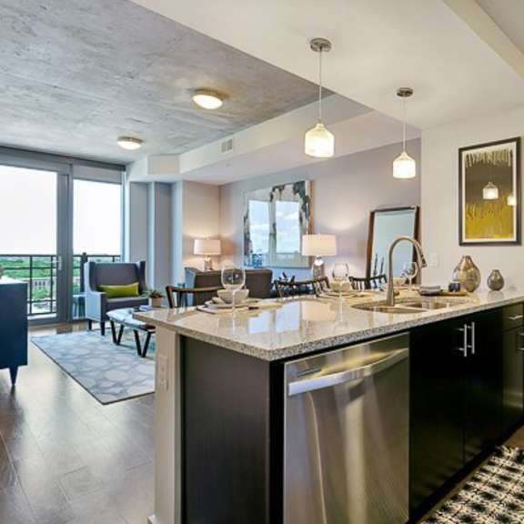 a modern kitchen with island here at victory place featuring dark cabinets, stainless steel appliances, pendant lighting, bar seating, and an open layout flowing into a bright living area with large windows.