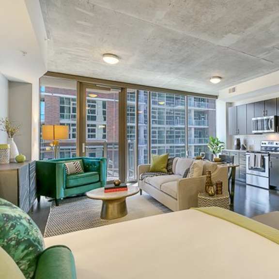 a bright open studio apartment here at victory place with a full modern kitchen, dark cabinets, stainless steel appliances, a cozy living area, green accent furniture, and large floor-to-ceiling windows with city views.