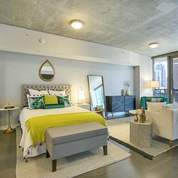 a stylish open studio bedroom and living area here at victory place with a tufted bed, green accent bedding, modern furniture, dark wood flooring, and large windows bringing in natural city views.