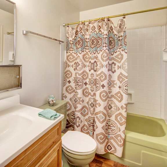 bright bathroom here at the village at calais apartments with wooden vanity, white sink, and a large mirror. features a green bathtub, patterned shower curtain, and window providing natural light.