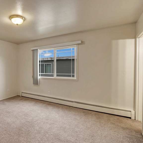 empty bedroom here at the village at calais apartments with beige carpet and off-white walls, featuring a large window with blinds and a ceiling light. natural light creates a bright, neutral ambiance.