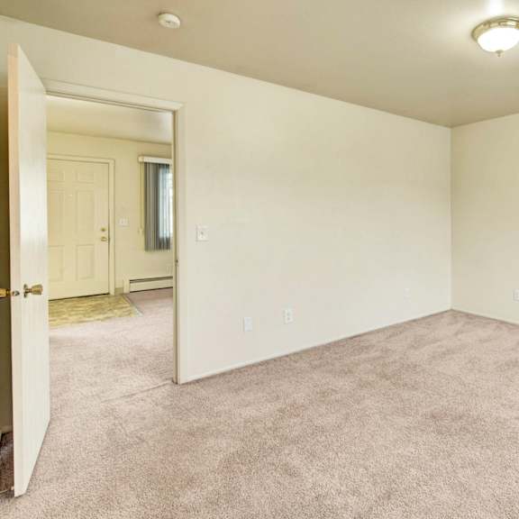 village at calaisempty beige room here at the village at calais apartments with carpeted floor, plain walls, and an overhead light fixture. an open door reveals an adjacent space with a window and a closed front door. minimalist and calm ambiance. apartments - livingroom