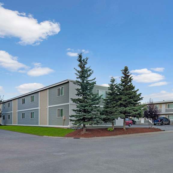 two-story gray village at calais apartment buildings with white trim, surrounded by lush trees and neatly manicured lawns under a bright blue sky with scattered clouds.