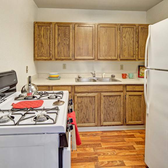 cozy kitchen here at the village at calais apartments with wooden cabinets, a gas stove, and white fridge. counter features a double sink and colorful mugs, conveying a warm, homely feel.