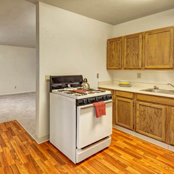 small kitchen the village at calais apartments with wooden cabinets, white stove, and wooden flooring. adjacent is a carpeted living area; both areas are empty, creating a neutral, simple tone.