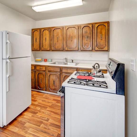 small kitchen here at the village at calais apartments with wood cabinets, white fridge, and gas stove. wooden floor, overhead light. cozy and simple, with a pot on the stove.