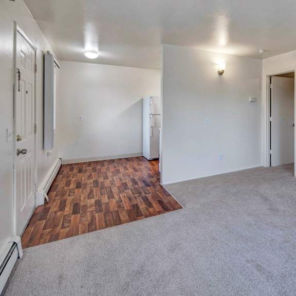 empty living room here at the village at calais apartments with light gray carpet and white walls, transitioning into a small kitchen area with wood-patterned flooring. minimalist and clean.