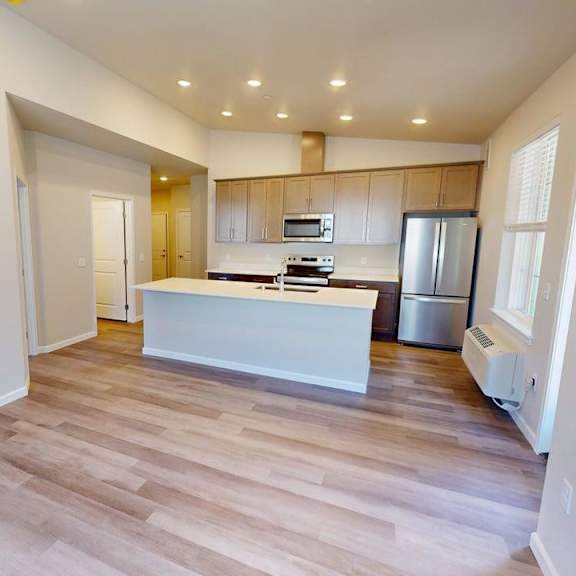 a spacious open living area here at autumn haus showcasing wide plank wood-style flooring, a bright open kitchen with a large white island, modern cabinetry, built-in appliances, and recessed ceiling lighting, with clean white walls, a window bringing in natural light, and an open layout ideal for both living and dining.