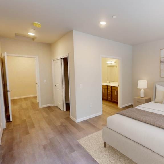 a serene primary bedroom here at autumn haus featuring wide plank wood-style flooring, a neutral upholstered bed with layered bedding, matching nightstands with lamps, and recessed ceiling lighting, with a private ensuite bathroom visible through the doorway, soft neutral walls, and a clean, spacious layout designed for comfort and relaxation.