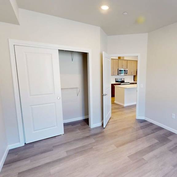 a bright unfurnished bedroom here at autumn haus featuring soft neutral walls, wood-style flooring, recessed ceiling lighting, and an open sliding-door closet with built-in shelving, with a doorway offering a clear view toward the kitchen area, creating a flexible and functional living space.