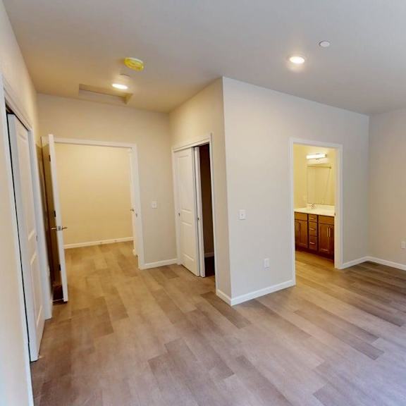 a spacious unfurnished bedroom here at autumn haus showcasing soft neutral walls, wood-style flooring, and recessed ceiling lighting, with an open layout that includes a sliding-door closet, a hallway leading toward the living areas, and a private bathroom entrance featuring a vanity and mirror, creating a clean and flexible space for personalization.