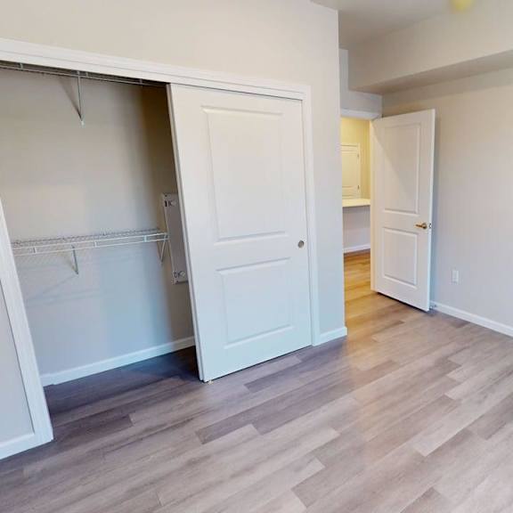 a versatile unfurnished bedroom here at autumn haus featuring soft neutral walls, wood-style flooring, and recessed ceiling lighting, with an open sliding-door closet offering wire shelving, ample wall space for furniture placement, and a doorway leading toward the hallway and adjacent living areas for a functional and flexible layout.