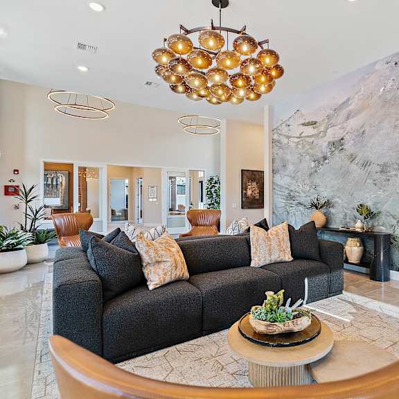 a stylish community lounge here at autumn haus featuring a dark upholstered sofa with patterned accent pillows, caramel-toned lounge chairs, a round coffee table with decorative greenery, polished flooring, a large abstract mural wall, statement globe pendant lighting, layered rugs, and an open layout that creates a warm and inviting space for relaxing or socializing.
