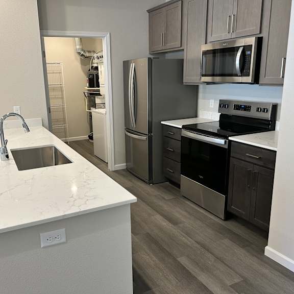 a modern kitchen here at autumn haus featuring a spacious white quartz-style island with an undermount sink, wide plank flooring, gray cabinetry, and sleek appliances, complemented by a pantry or utility room visible through an open doorway that adds extra storage and everyday convenience.