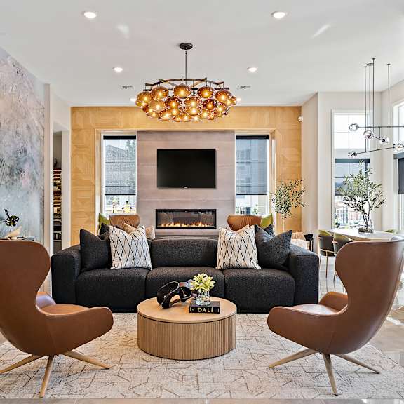 a modern community lounge here at autumn haus featuring a plush dark sofa with patterned accent pillows, sculptural caramel-toned lounge chairs, a round wood coffee table with decorative accents, a sleek fireplace with a mounted television, statement globe pendant lighting, large windows, and an abstract mural wall creating a warm and sophisticated gathering space.