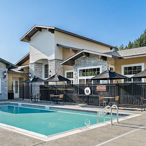 a resort-style outdoor pool here at autumn haus featuring a rectangular swimming pool with metal handrails, textured concrete decking, shaded seating areas with black umbrellas and café tables, safety fencing with mounted signage, and a stone-accented clubhouse façade with large windows set beneath a clear blue sky.