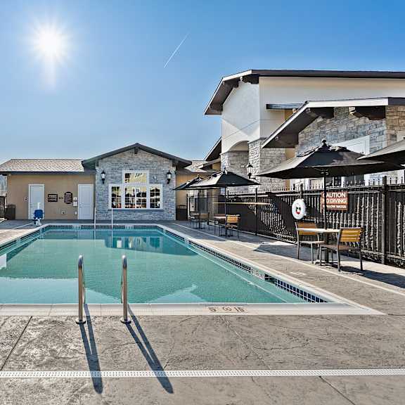 a sunlit resort-style pool here at autumn haus featuring a rectangular swimming pool with centered metal handrails, textured concrete decking, clearly marked depth indicators, and surrounding lounge seating with black umbrellas, all enclosed by safety fencing and framed by a stone-accented clubhouse and forested hillside views under a clear blue sky.