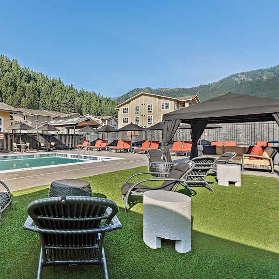 a relaxing outdoor pool lounge here at autumn haus featuring a sparkling rectangular swimming pool bordered by textured concrete decking, cushioned chaise lounges with orange accents, woven patio chairs on green turf, shaded cabanas, and safety fencing, all set against stone-accented buildings and forested hillside views beneath a clear blue sky.