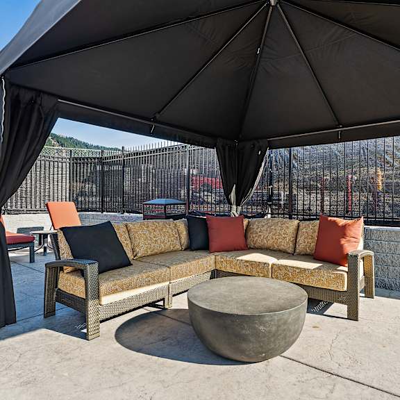 a shaded outdoor cabana lounge here at autumn haus featuring a covered pavilion with dark drapery, a woven sectional sofa with warm neutral and rust accent pillows, a round concrete coffee table, and nearby chaise lounges on textured concrete decking, all enclosed by fencing with hillside and mountain views in the background.