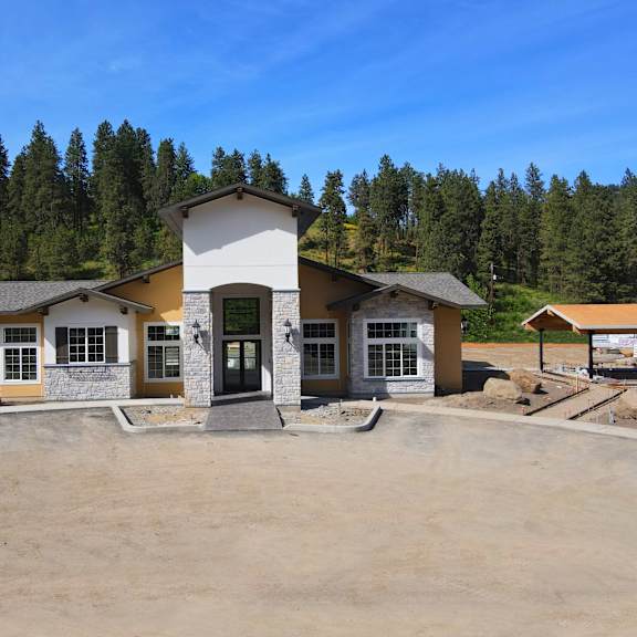 a welcoming clubhouse exterior here at autumn haus showcasing a modern lodge-style building with stone accents, warm-toned siding, large grid windows, and a central covered entry, set against a backdrop of tree-lined hills and open sky with surrounding walkways and landscaped grounds.