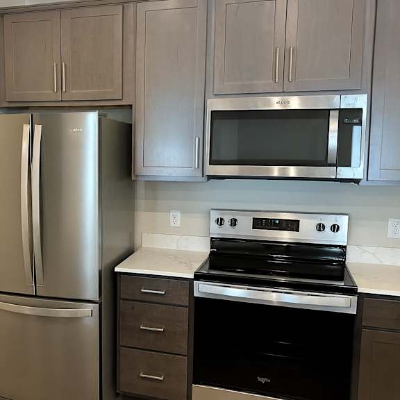a modern kitchen here at autumn haus showcasing gray shaker-style cabinetry, white quartz-style countertops, and sleek appliances, with a full-size refrigerator and range framed by upper cabinets, while an open doorway reveals a convenient in-home laundry area that adds functionality and everyday ease to the space.