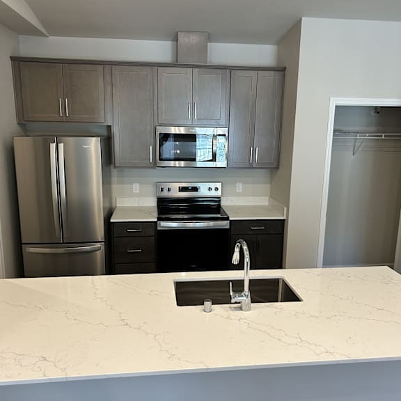 a contemporary kitchen here at autumn haus featuring gray shaker-style cabinets, a wide white quartz-style island with an undermount sink and modern faucet, and sleek stainless-style appliances, with ample counter space, clean-lined finishes, and an open layout that creates a functional and inviting cooking and gathering area.