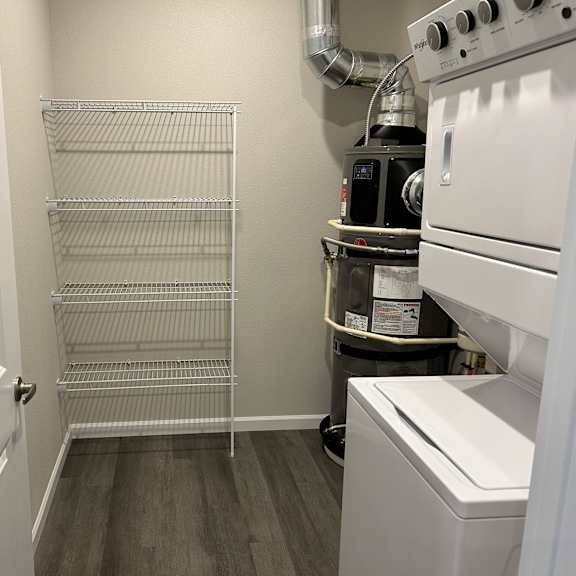 a functional laundry and storage area here at autumn haus featuring a full-size stacked washer and dryer, durable wood-style flooring, built-in wire shelving for organization, neutral painted walls, and an efficient layout that provides convenient space for laundry tasks and everyday household storage.