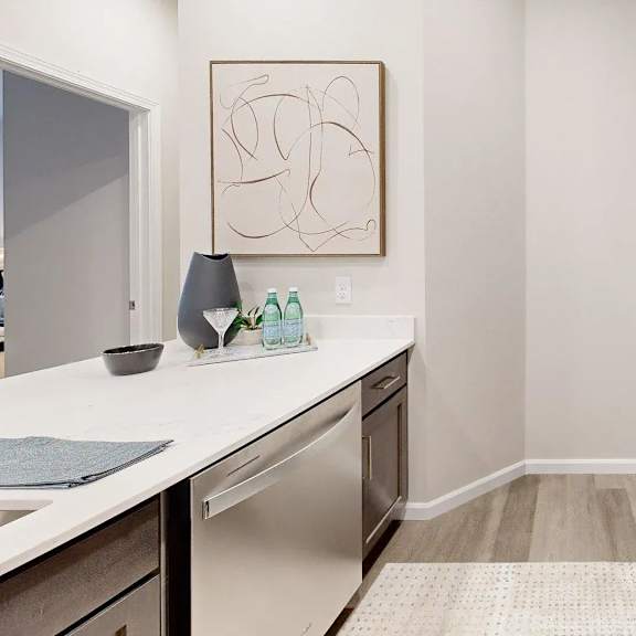a sleek galley-style kitchen here at autumn haus featuring smooth white countertops, warm wood cabinetry with modern hardware, a built-in dishwasher, microwave, and oven, open counter space with decorative accents, wood-style flooring, and a clean, bright layout designed for efficient cooking and everyday living.